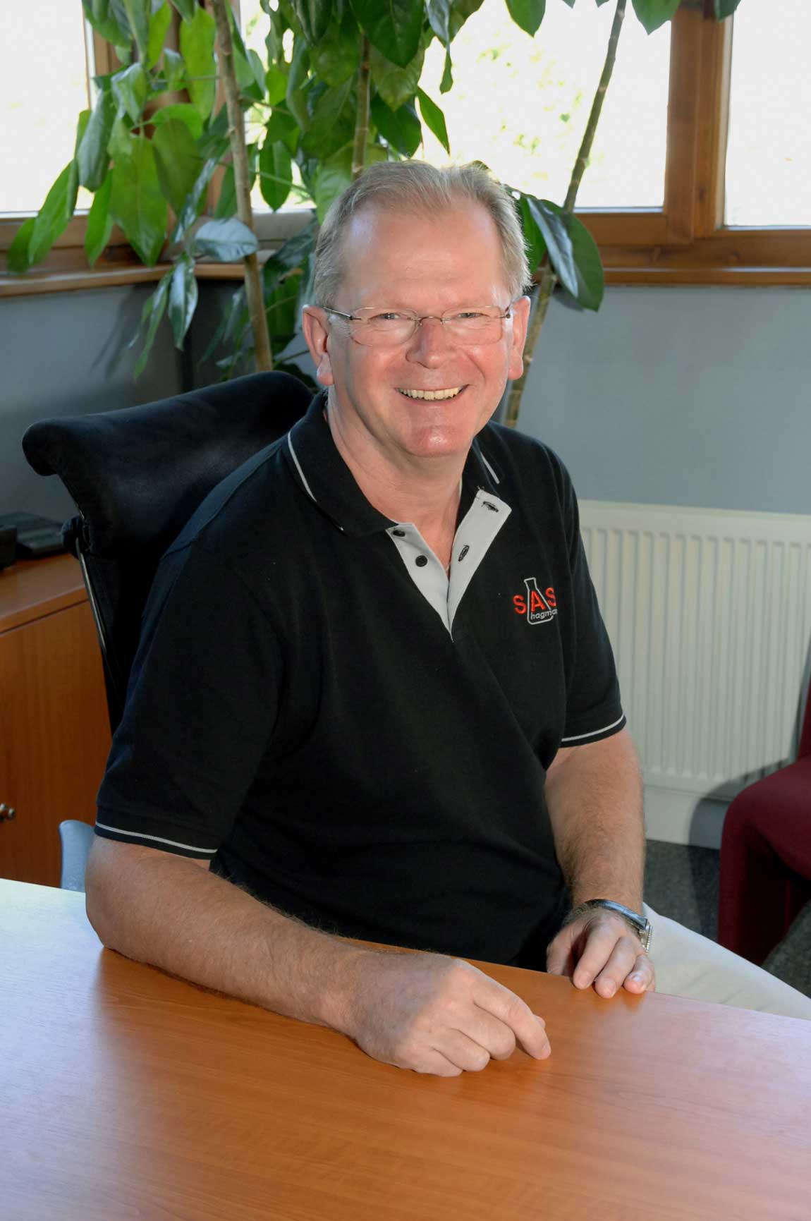 Person mit schwarzen Polo-Shirt sitzt lächelnd auf seinem Bürostühl und leht sich auf sein Bürotisch