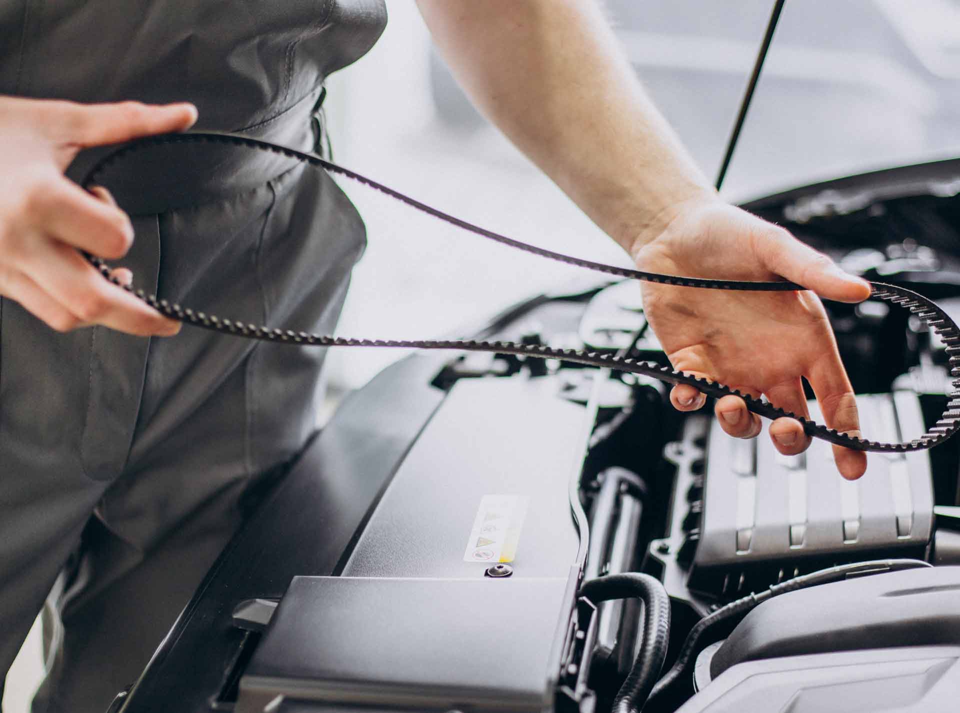 Eine Person hantiert mit einem Keilriemen über einem geöffneten Fahrzeugmotor, offenbar bei einer Inspektion oder Wartung in einer Werkstattumgebung.