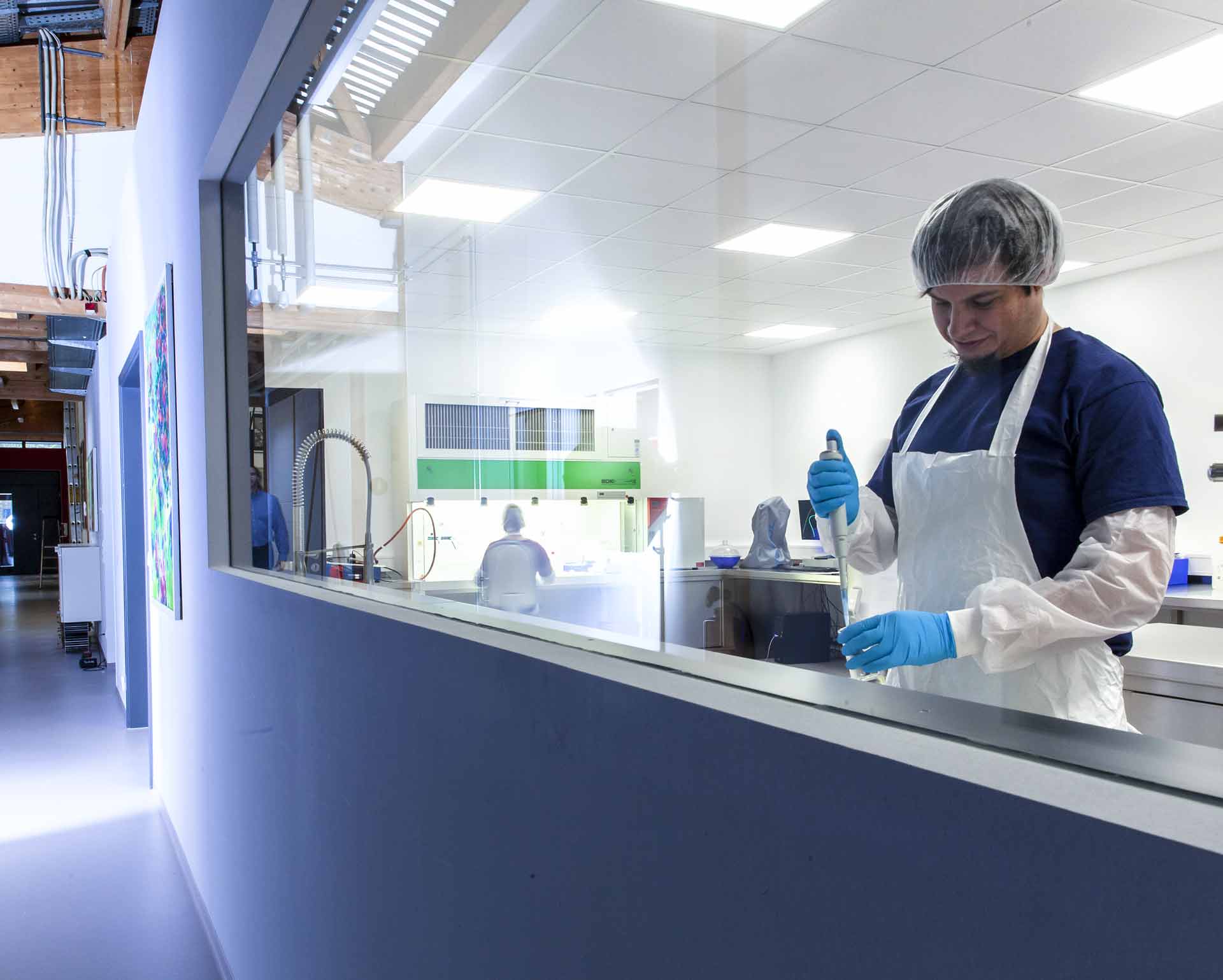 Person in Schutzkleidung und Haarnetz pipettiert Flüssigkeit in einem modernen Labor, während eine andere Person im Hintergrund an einem Tisch arbeitet.