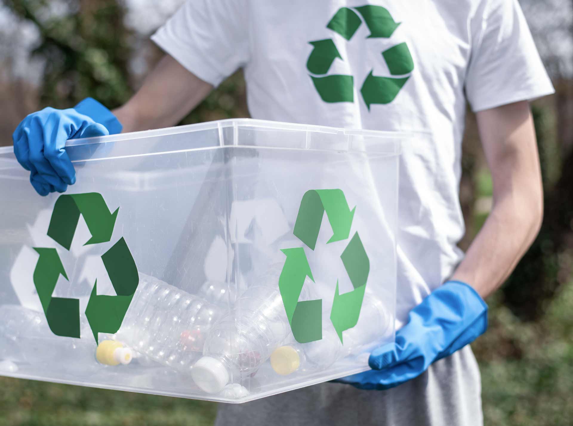 Person hält transparente Box mit Plastikflaschen, trägt recycelbarem Symbol auf Kleidung im Freien. Box und T-Shirt zeigen grünes Recycling-Symbol, blaue Handschuhe runden das Bild ab.