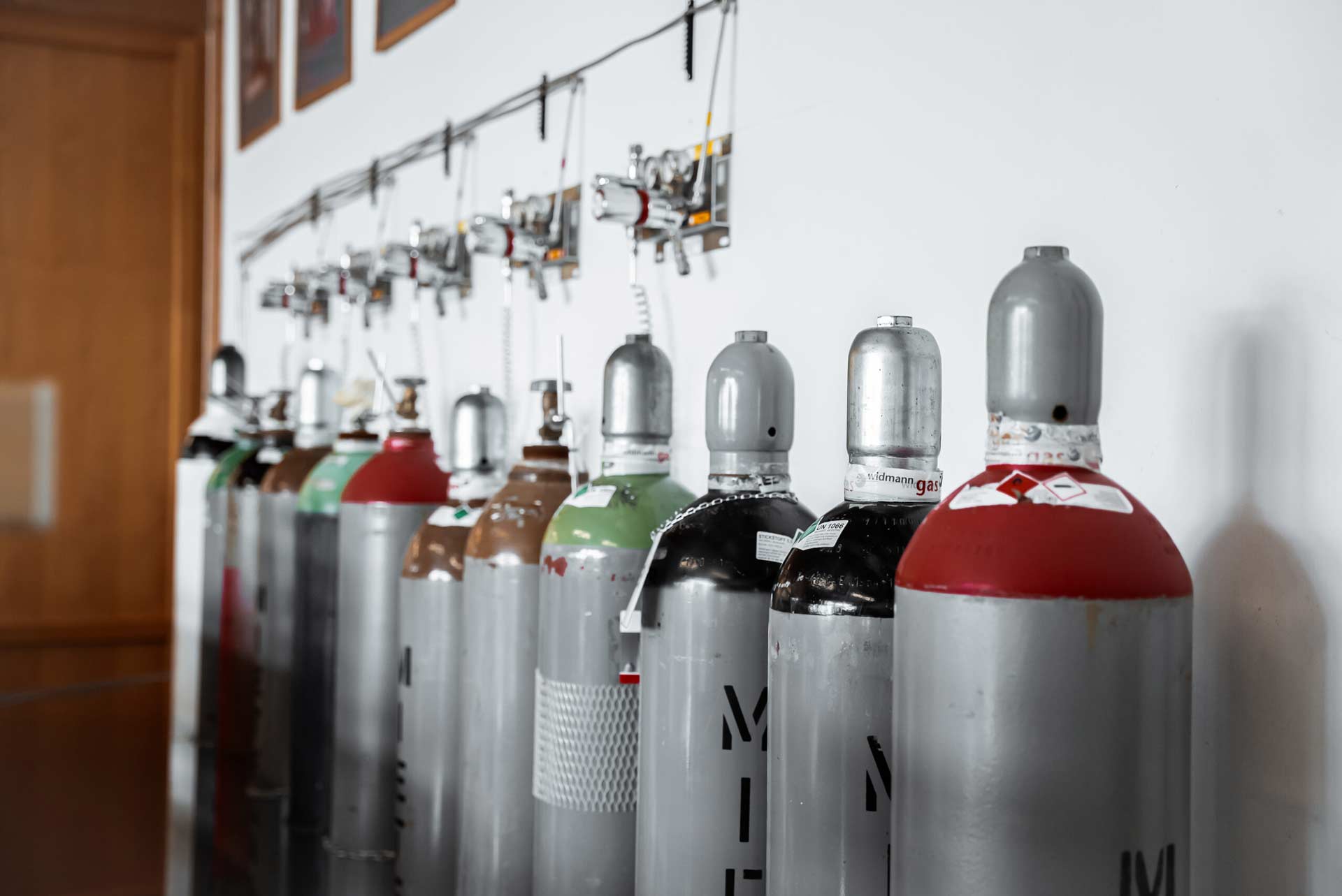 Mehrere Gasflaschen stehen in einer Reihe an der Wand, verbunden mit Ventilen und Schläuchen. Die Flaschen haben unterschiedliche Farben und sind deutlich beschriftet.