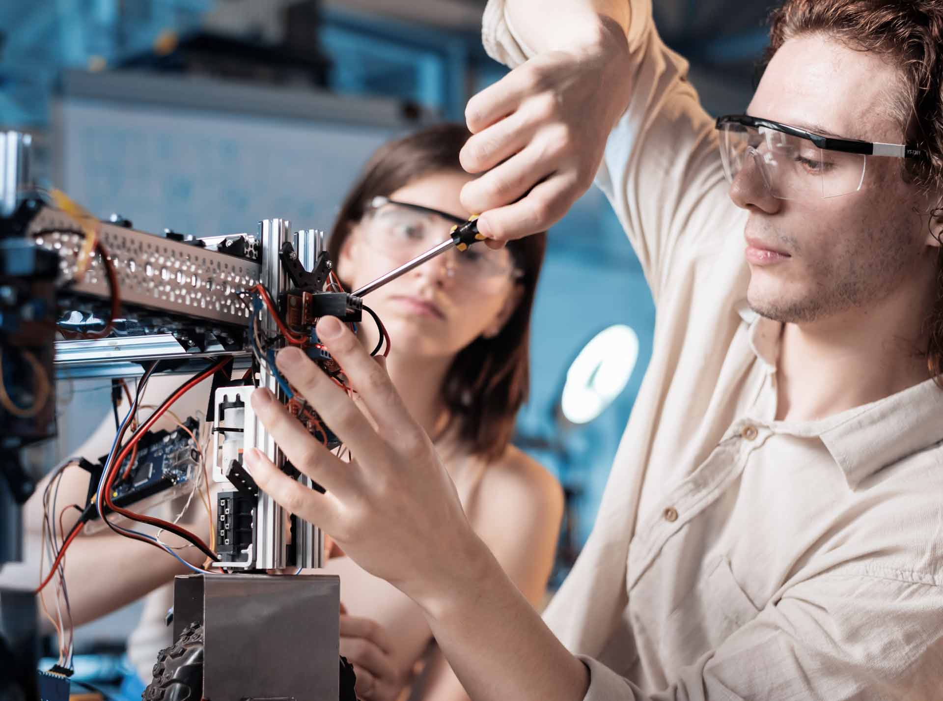 Zwei Personen arbeiten an einem technischen Gerät mit elektronischen Komponenten, Schraubendreher in Hand, tragen Schutzbrillen, in einer Werkstattumgebung.