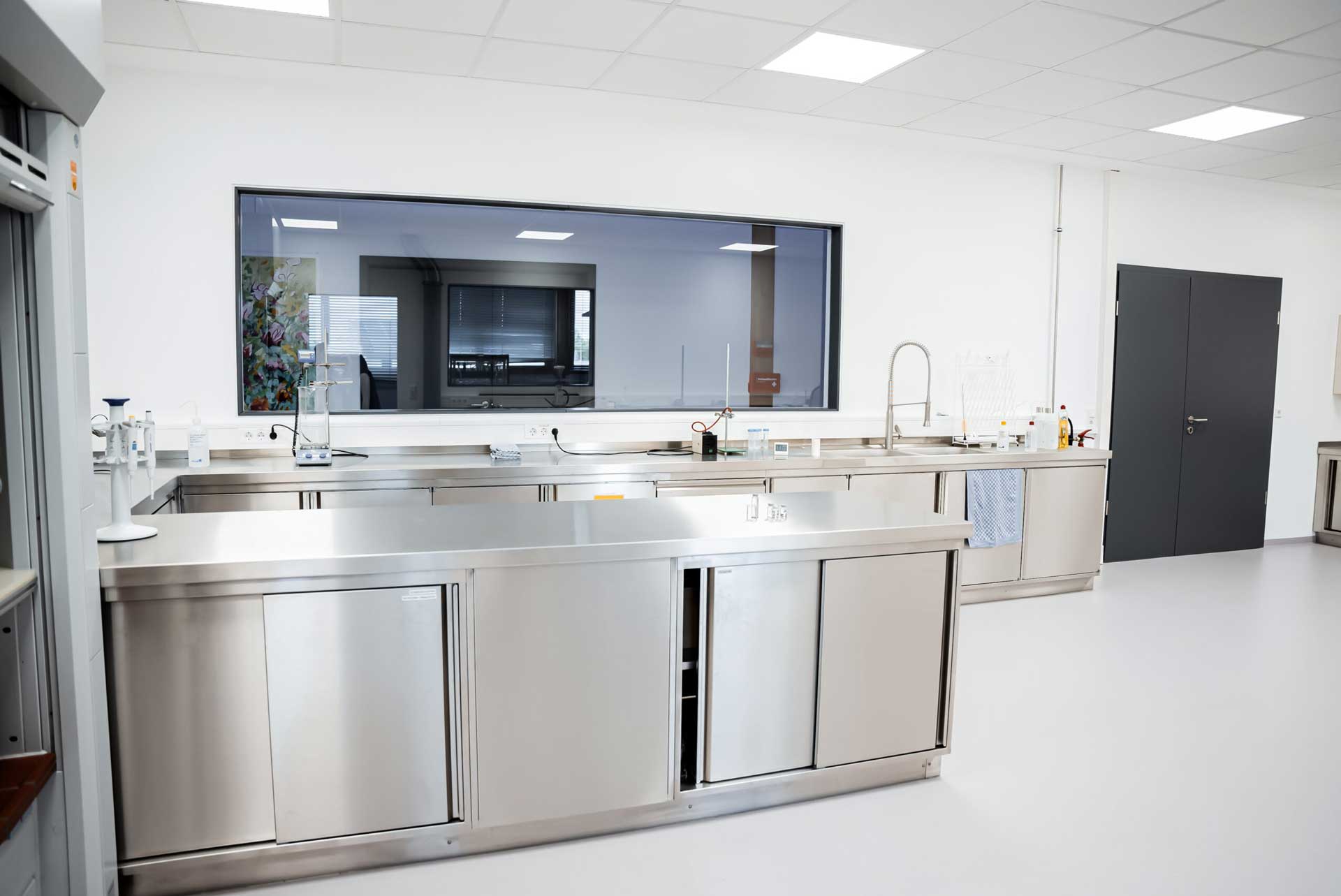 Edelstahl-Arbeitsbereich – sauber und leer – moderner Laborraum mit großen Fenstern und weißen Wänden, ausgestattet mit Laborgeräten und einem Wasserhahn, ohne Personen oder sichtbare Aktivitäten.