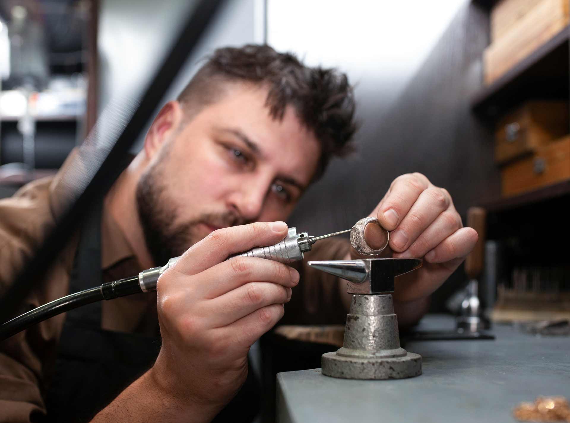 Ein Mann bearbeitet sorgfältig einen Ring mit einem kleinen Bohrwerkzeug in einer Werkstatt, umrandet von Werkzeugen und Holzschränken.