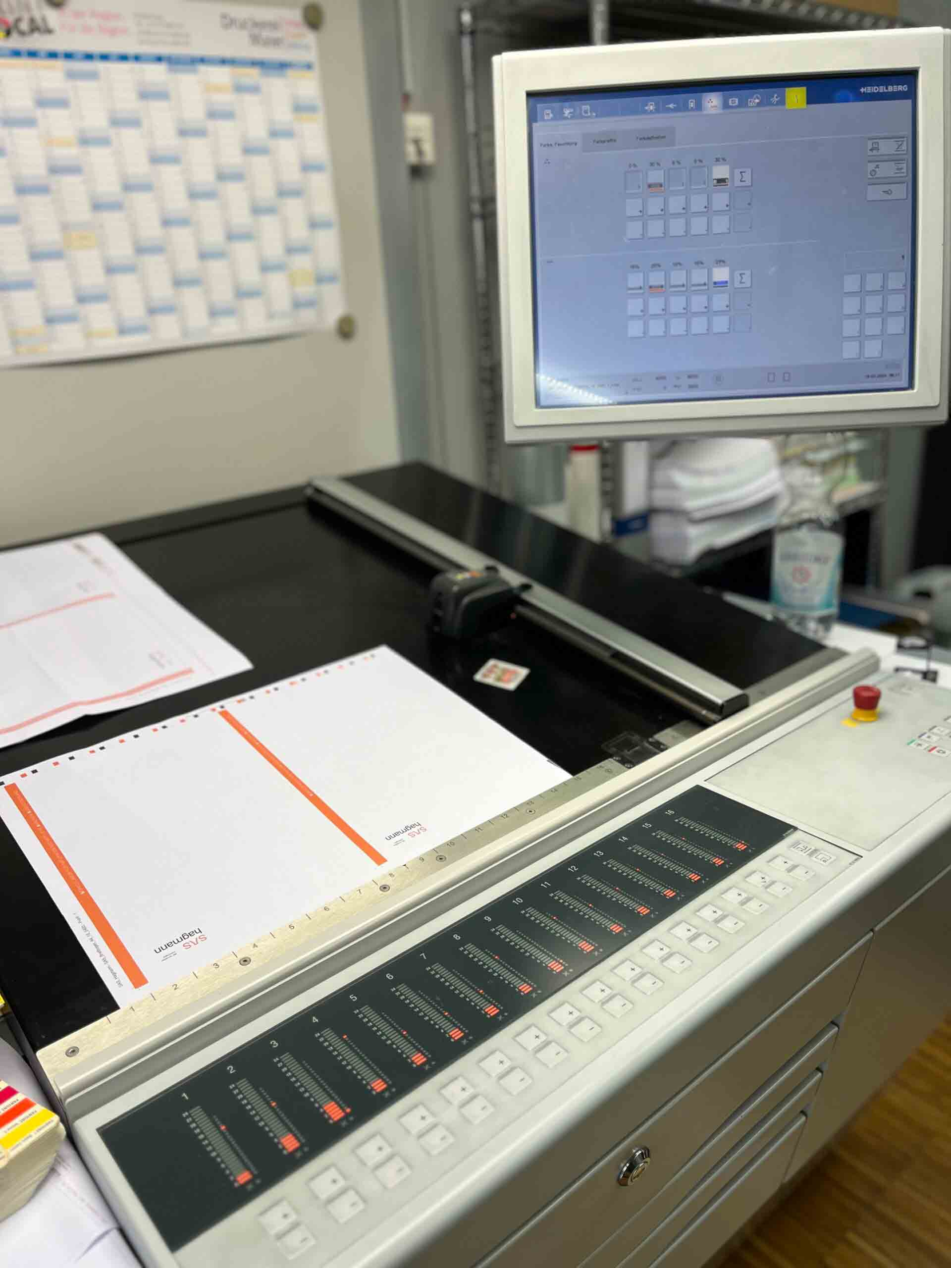 Monitor zeigt Druckkontrollsoftware, daneben Druckbögen mit Farbtabelle; Steuerpult mit Reglern und Knöpfen, im Druckereiumfeld. Text auf Bögen: „Ingenium“ und „Ligneum“.