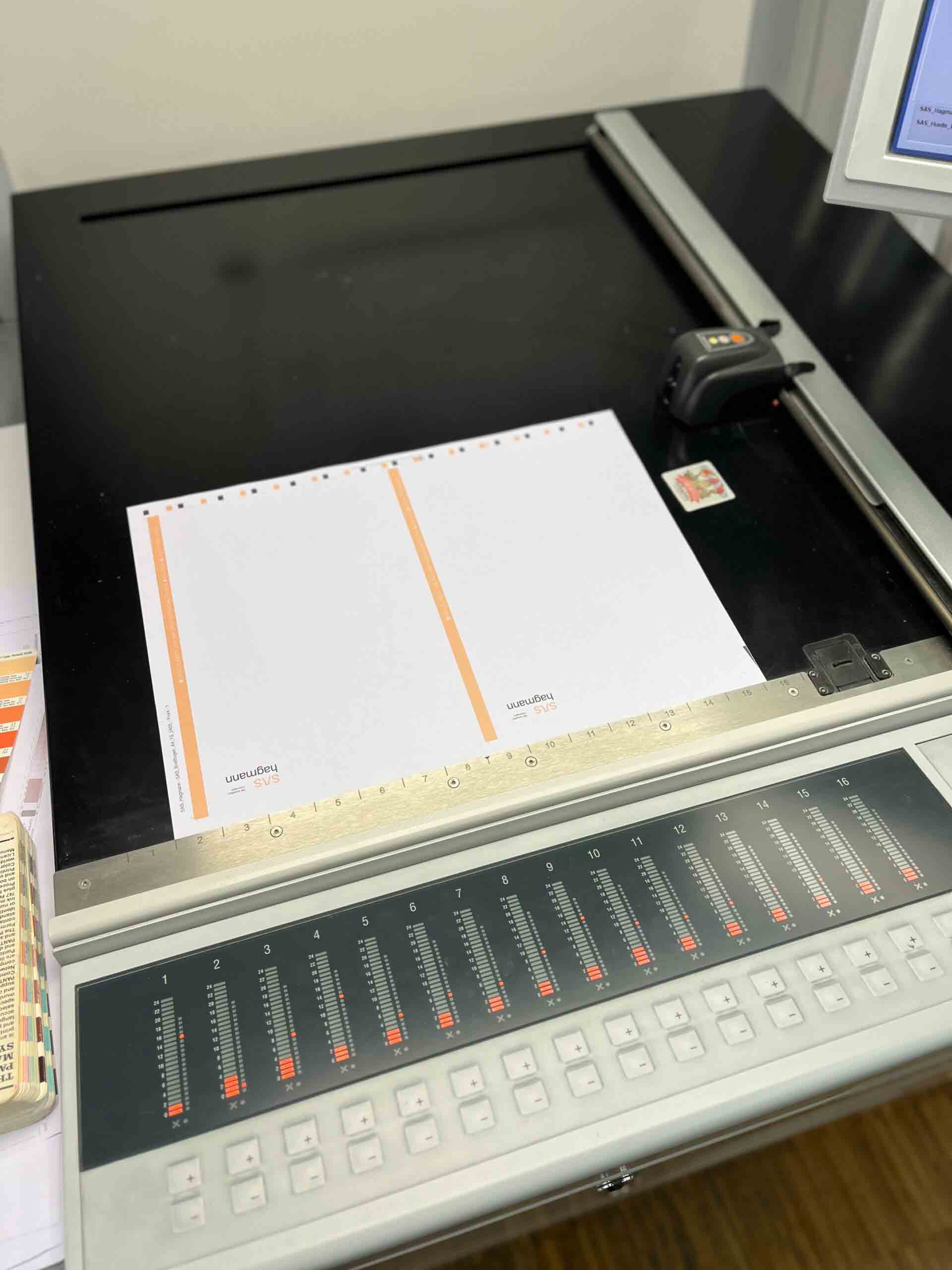 Ein Kontrollpult mit Schiebereglern und Tasten, vor einem Scanner. Darauf liegen zwei leere, weiß-orange Seiten von SLS Spangenberg.