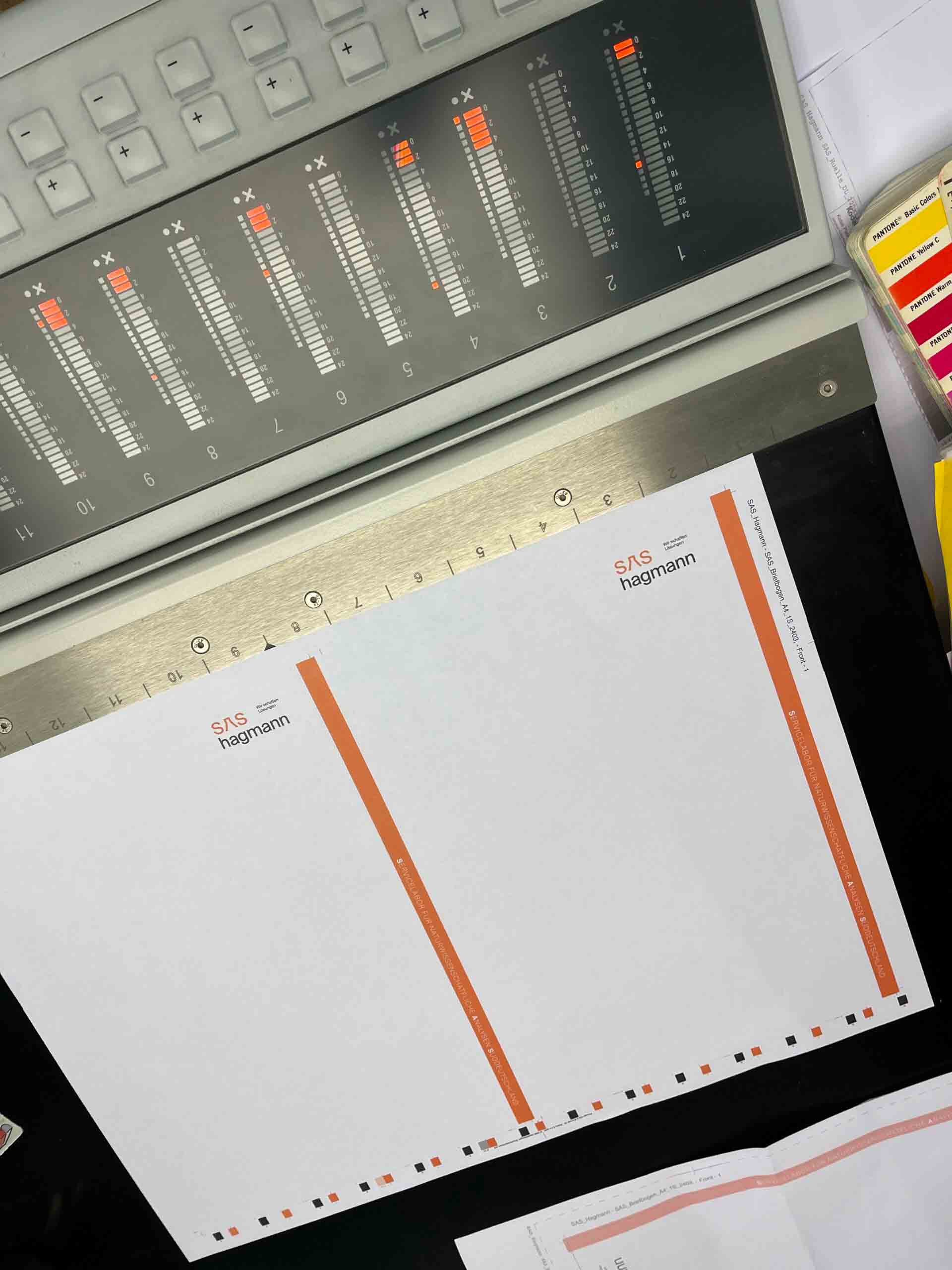 Ein Steuerpult mit Schiebereglern und Tasten. Darunter liegt ein Papierbogen mit dem Text „SAS hagmann“ und einem orangen Streifenmuster. Daneben Farbmusterkarten.