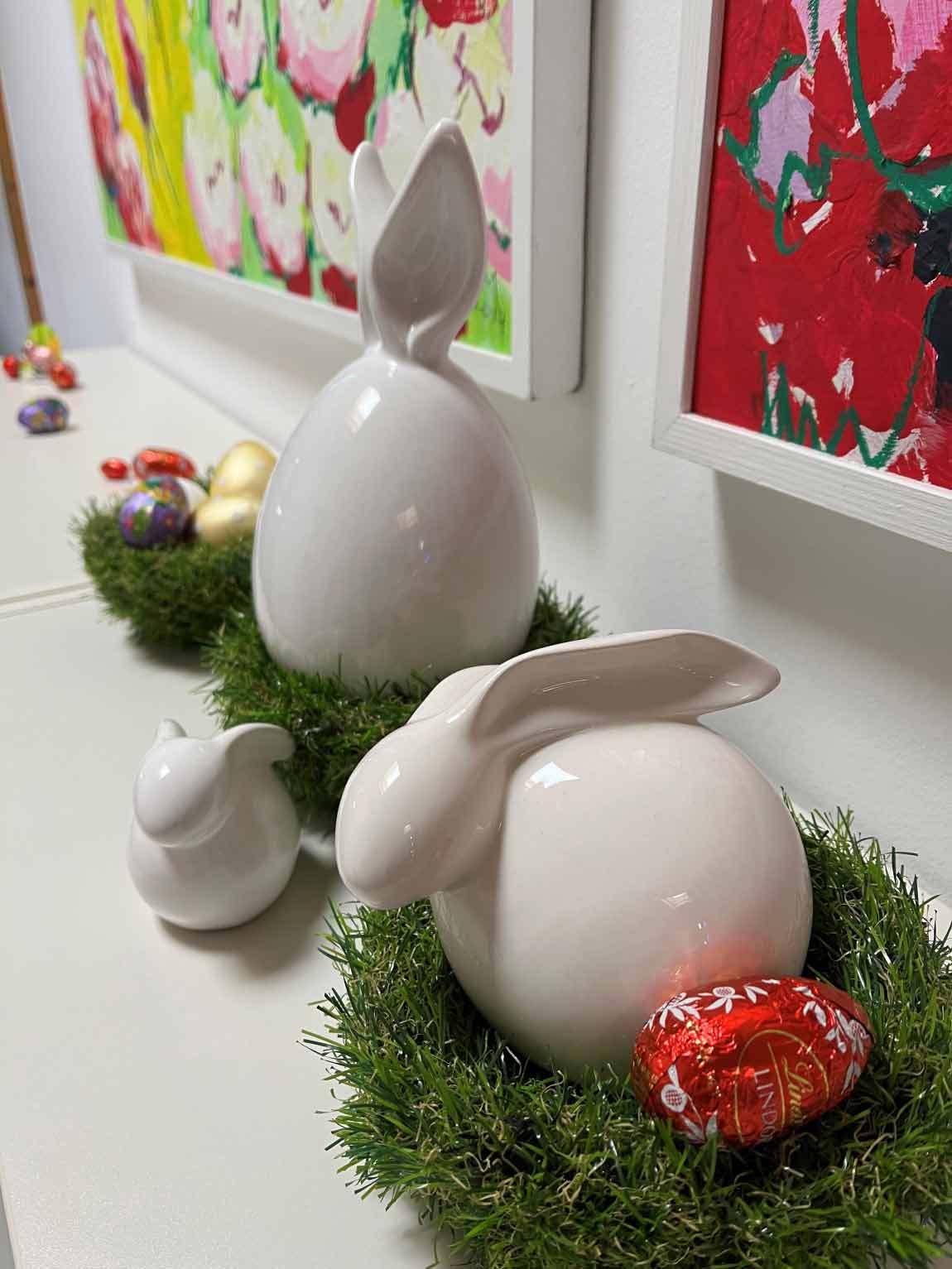 Keramik-Osterhasen stehen dekorativ auf künstlichem Gras neben bemalten Eiern und einem Lindt-Schokoladenei, vor leuchtenden Blumenmalereien an der Wand.