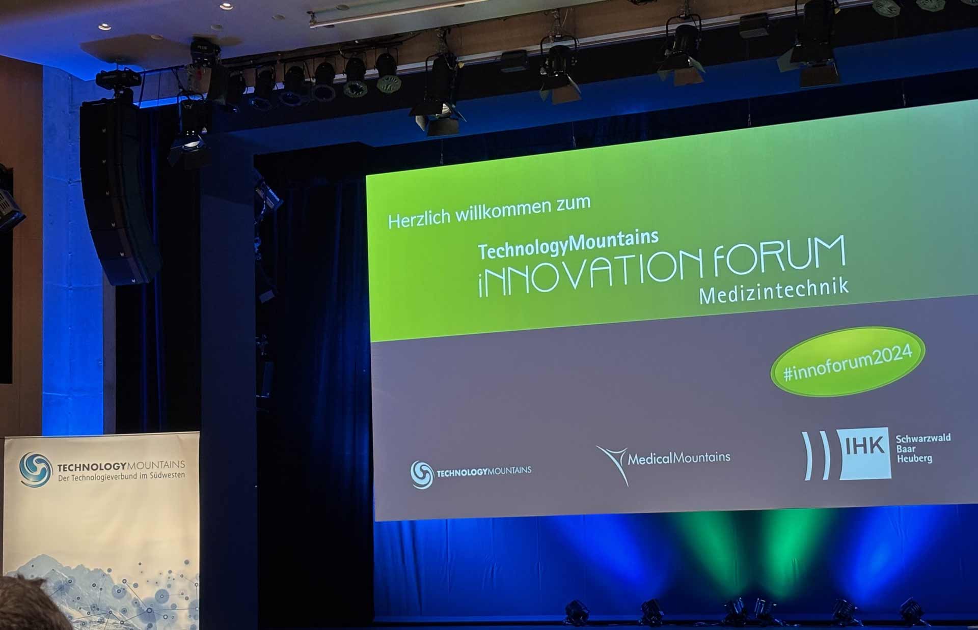 Bühne mit blau-grün beleuchtetem Display zeigt Text: „Herzlich willkommen zum TechnologyMountains Innovation Forum Medizintechnik.“ Logos von TechnologyMountains, MedicalMountains, IHK. Banner links mit TechnologyMountains-Logo.
