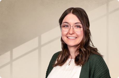 Eine Frau mit Brille lächelt in die Kamera. Sie trägt einen grünen Cardigan und steht vor einer Wand mit Schatten von Fenstern. - Anna Echle