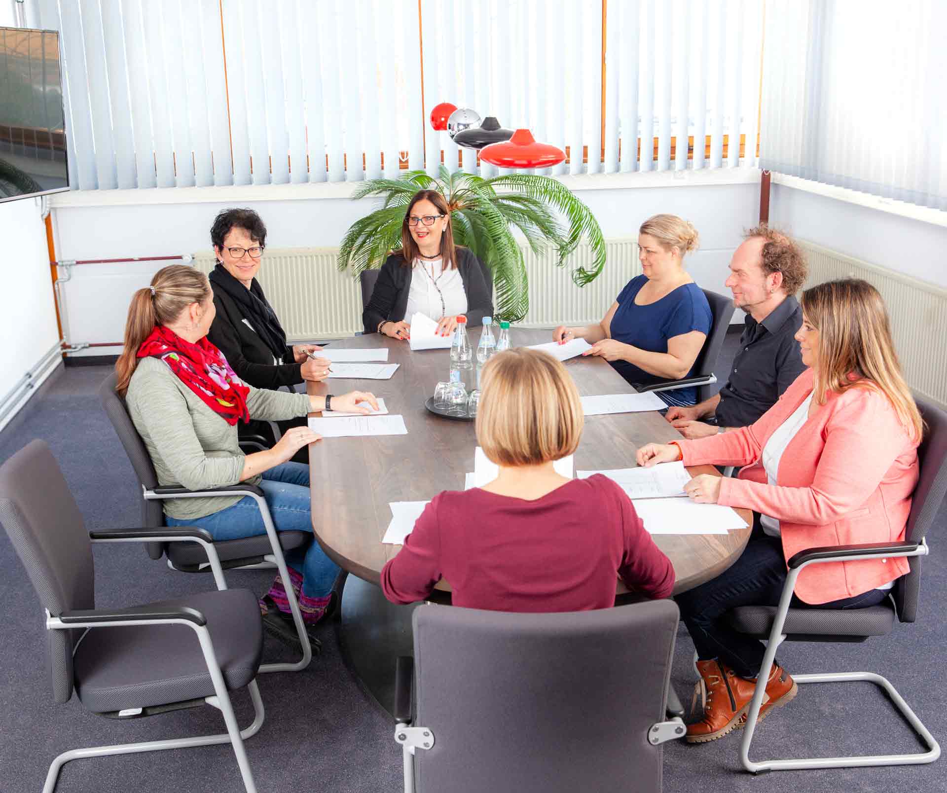 Eine Gruppe von sieben Personen sitzt um einen rechteckigen Tisch in einem Konferenzraum und diskutiert. Sie halten Papierblätter, Wasserflaschen stehen auf dem Tisch.