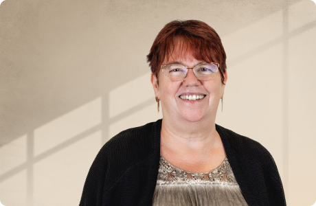 Eine lächelnde Person steht vor einem Hintergrund mit weich einfallenden Schattenlinien, trägt eine Brille und ein gemustertes Oberteil. - Brigitte Herberg Justen