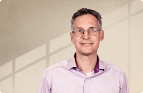 Ein Mann mit Brille lächelt in die Kamera. Er trägt ein lila Hemd. Im Hintergrund sind verschwommene Fensterrahmen zu sehen. - Dr. Bernd Görlach