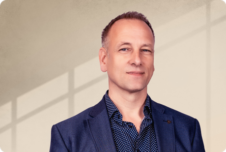 Eine Person in einem blauen Anzug steht vor einer beigen Wand mit Schattenmuster, blickt selbstbewusst in die Kamera. - Dr. Nikolas Pietrzik