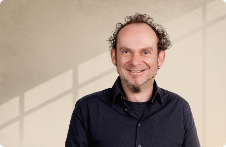 Ein Mann mit lockigem Haar und gepflegtem Bart lächelt in die Kamera; er trägt ein dunkles Hemd vor einem hellen Hintergrund mit Schattenlinien. - Dr. Peter Tremmel