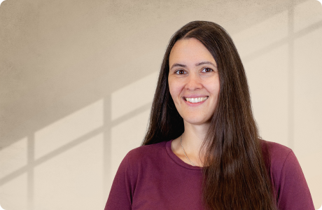 Eine Person mit langen dunklen Haaren lächelt in die Kamera. Sie trägt ein bordeauxrotes Oberteil. Im Hintergrund ist eine helle Wand mit Schatten eines Fensters erkennbar. - Kerstin Scherzinger