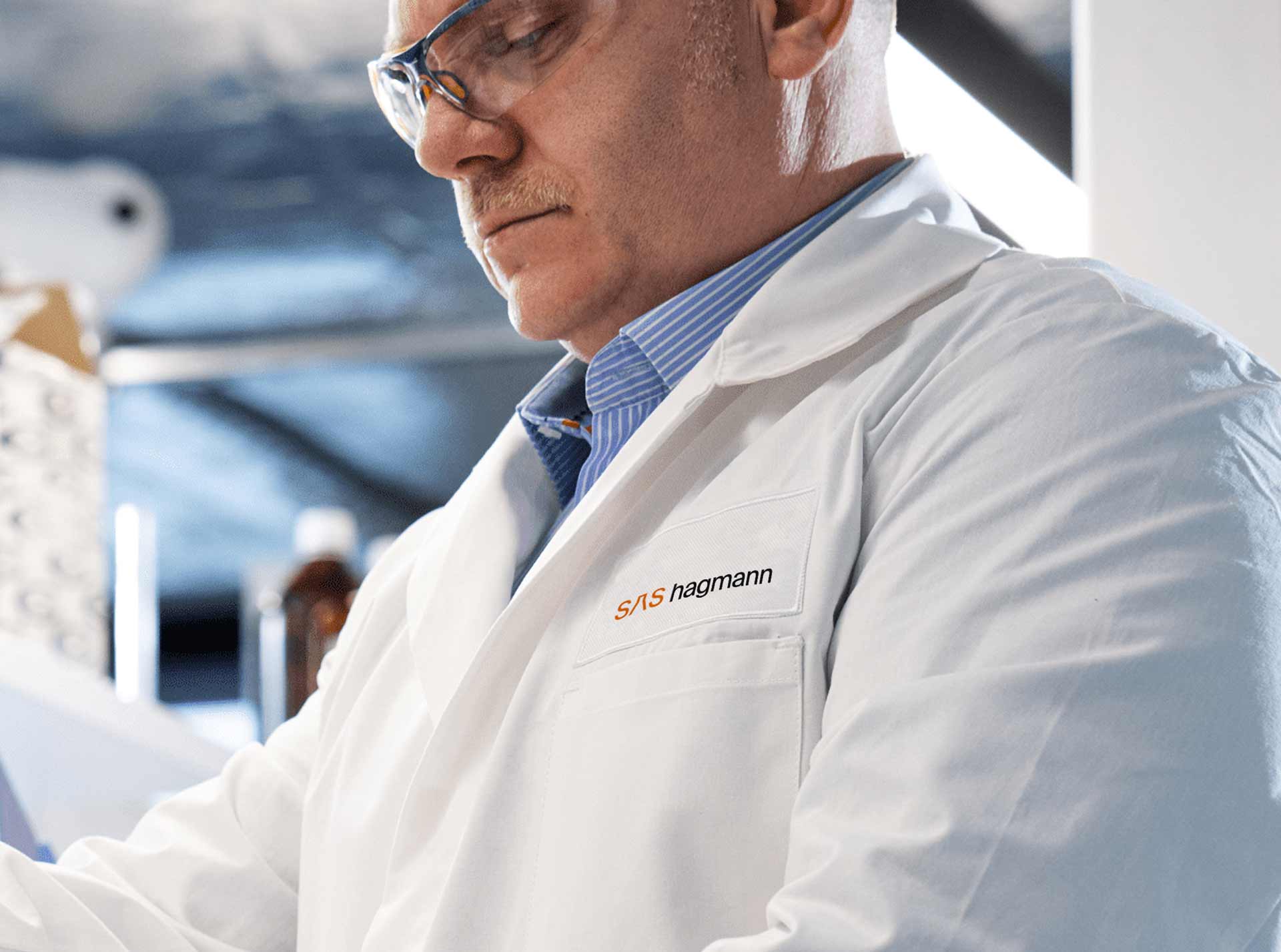 Mann in weißem Labor-Kittel mit Aufschrift "sas hagmann" arbeitet konzentriert, trägt Schutzbrille. Hintergrund zeigt Laborumgebung mit Flaschen und Geräten.