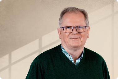 Eine Person mit Brille trägt einen grünen Pullover und ein gestreiftes Hemd, lächelt und steht vor einer Wand mit Licht- und Schattenmustern. - Manfred Hagmann