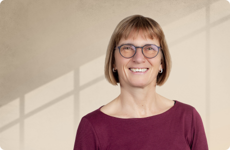 Person lächelt, steht vor einer beigen Wand mit Schatten in Rechteckmuster. Sie trägt eine Brille und ein dunkelrotes Oberteil. - Marlies Eisele
