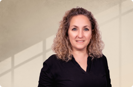 Eine Person mit lockigem Haar steht vor einem sanft beleuchteten Hintergrund. Sie trägt ein dunkles Oberteil und lächelt leicht in die Kamera. Schattenlinien sind sichtbar. - Mercedes Ramirez