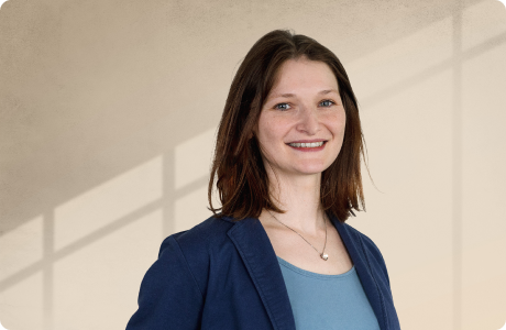 Eine lächelnde Person in einem blauen Blazer steht vor einem hellen, schattierten Hintergrund. Sie trägt eine Halskette und ein blaues Oberteil. - Monika Breithaupt