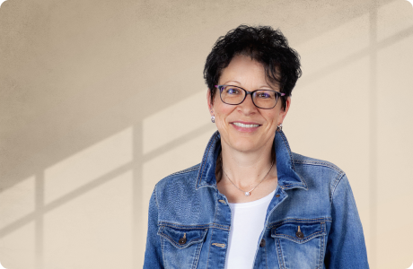 Eine Person mit Brille und Jeansjacke lächelt in die Kamera, vor einem beige Hintergrund mit Schattenmuster. - Nicole Kleffner