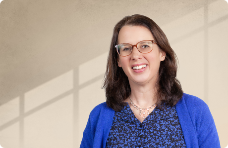 Eine lächelnde Person mit Brille trägt eine blaue Bluse und einen blauen Pullover, steht vor einem Fenster mit Schattenmuster. - Phillippa Landenberger