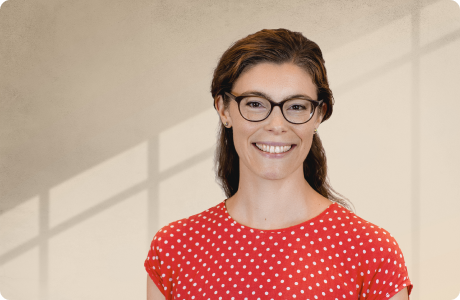 Eine Frau mit Brille lächelt und trägt ein rotes, gepunktetes Kleid. Sie steht vor einem neutralen Hintergrund mit Schattenlinien. - Rebecca Bauer