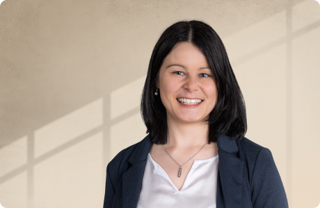 Eine lächelnde Frau mit dunklem, schulterlangem Haar trägt Bluse und Blazer vor neutralem Hintergrund mit Schattenmuster. - Selina Steinhilber