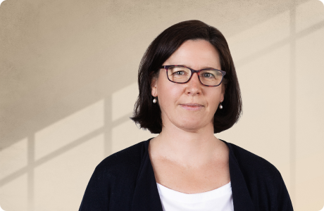 Eine Frau mit dunklem Haar und Brille steht vor einem beigen Hintergrund mit Schatten von Fensterrahmen. Sie trägt eine weiße Bluse und eine schwarze Jacke. - Solveig Schneiderhan