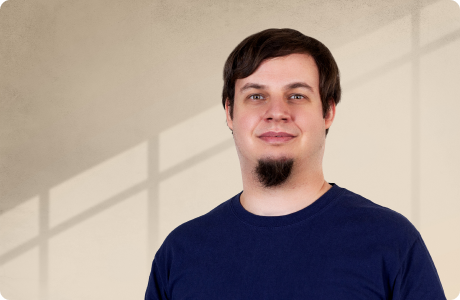 Eine lächelnde Person steht vor einem beigefarbenen Hintergrund mit Schattenlinien. Sie trägt ein dunkelblaues T-Shirt und hat kurzes, dunkles Haar. - Sven Rybka