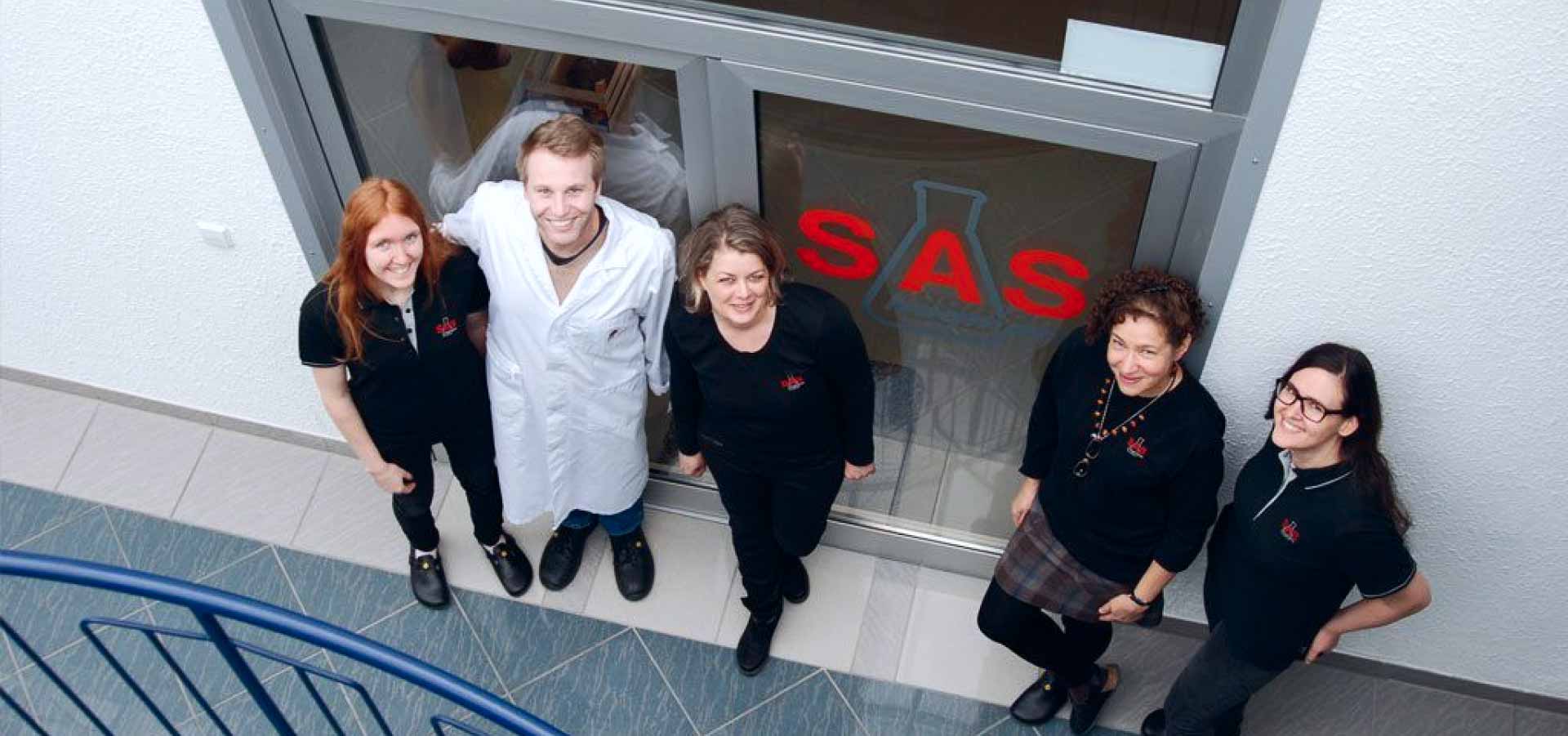 Fünf Personen lächeln in die Kamera, stehend vor einer Glastür mit rotem "SAS"-Logo, in einem Innenraum mit blauen Geländer und grauen Fliesen.