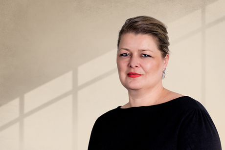 Eine Frau steht vor einem hellen Hintergrund mit Schattenlinien, trägt ein schwarzes Oberteil und schaut in die Kamera. - Tina Schwering