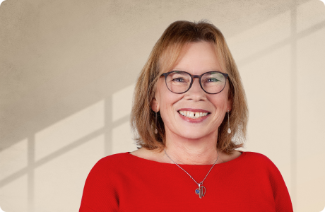 Eine Frau in rotem Oberteil lächelt direkt in die Kamera. Sie trägt eine Brille, schmales Schmuckstück. Der Hintergrund zeigt eine beige Wand mit Schattenlinien. - Wilma Mast